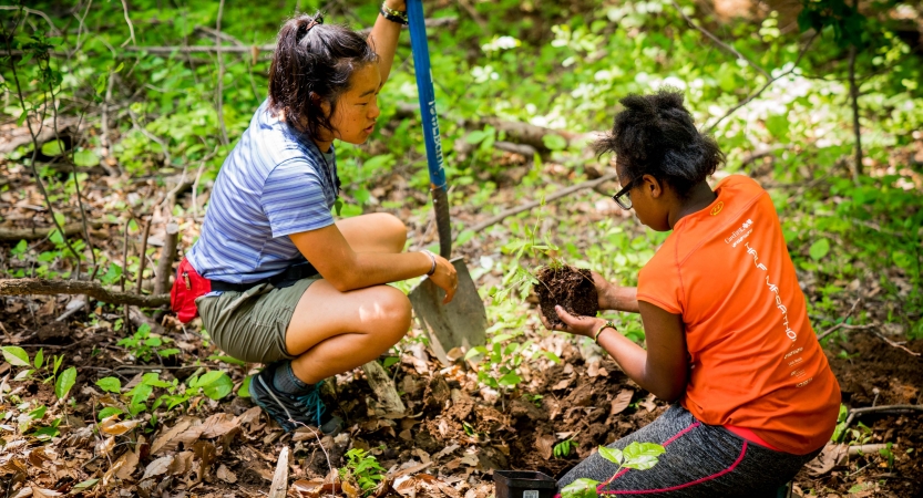 One person holds a shovel and another holds a mound of dirt. 